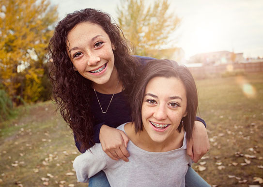 two smiling girls with braces, one riding piggyback on the other in a field