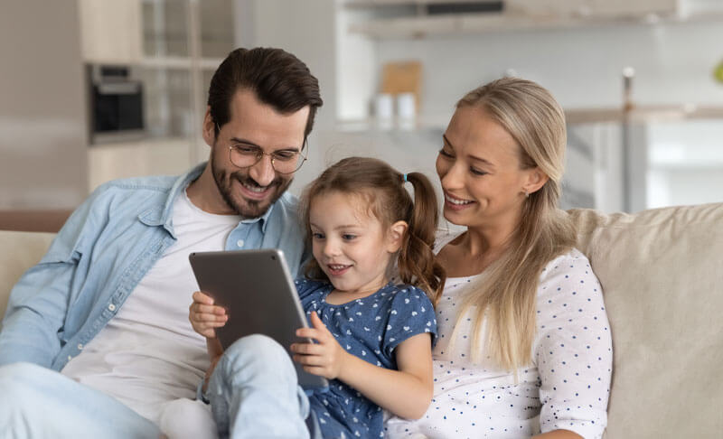 father and mother sitting on a couch with their young daughter, who is holding a tablet